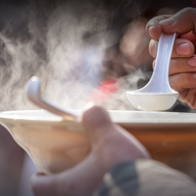 A hand holding a white spoon above a steaming bowl of soup.