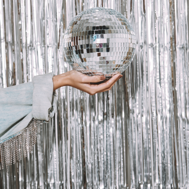 A hand holding a small disco ball in front of shiny silver curtains.